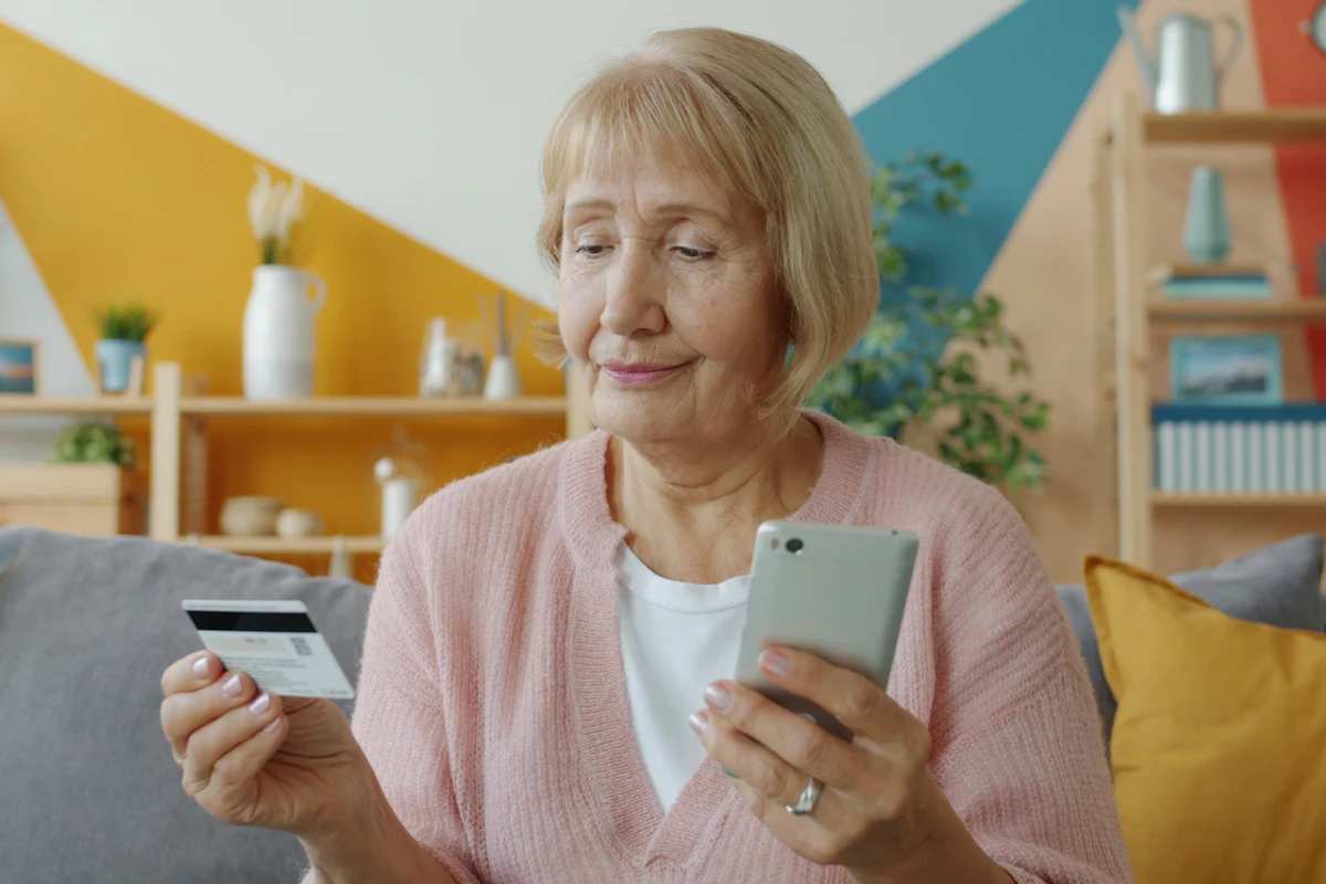 An older woman checks her phone while holding a bank card.